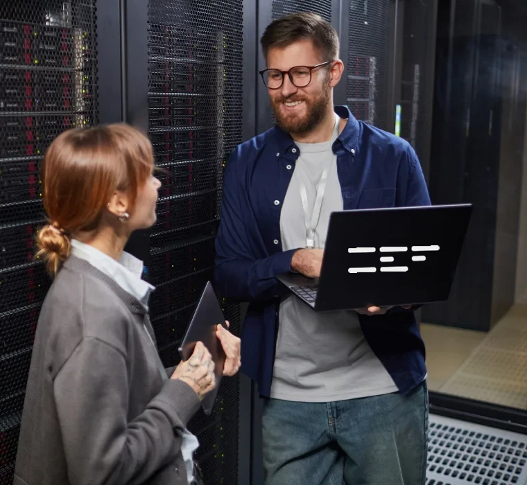 Two engineers talking beside server racks in a data centre, one holding an open laptop.